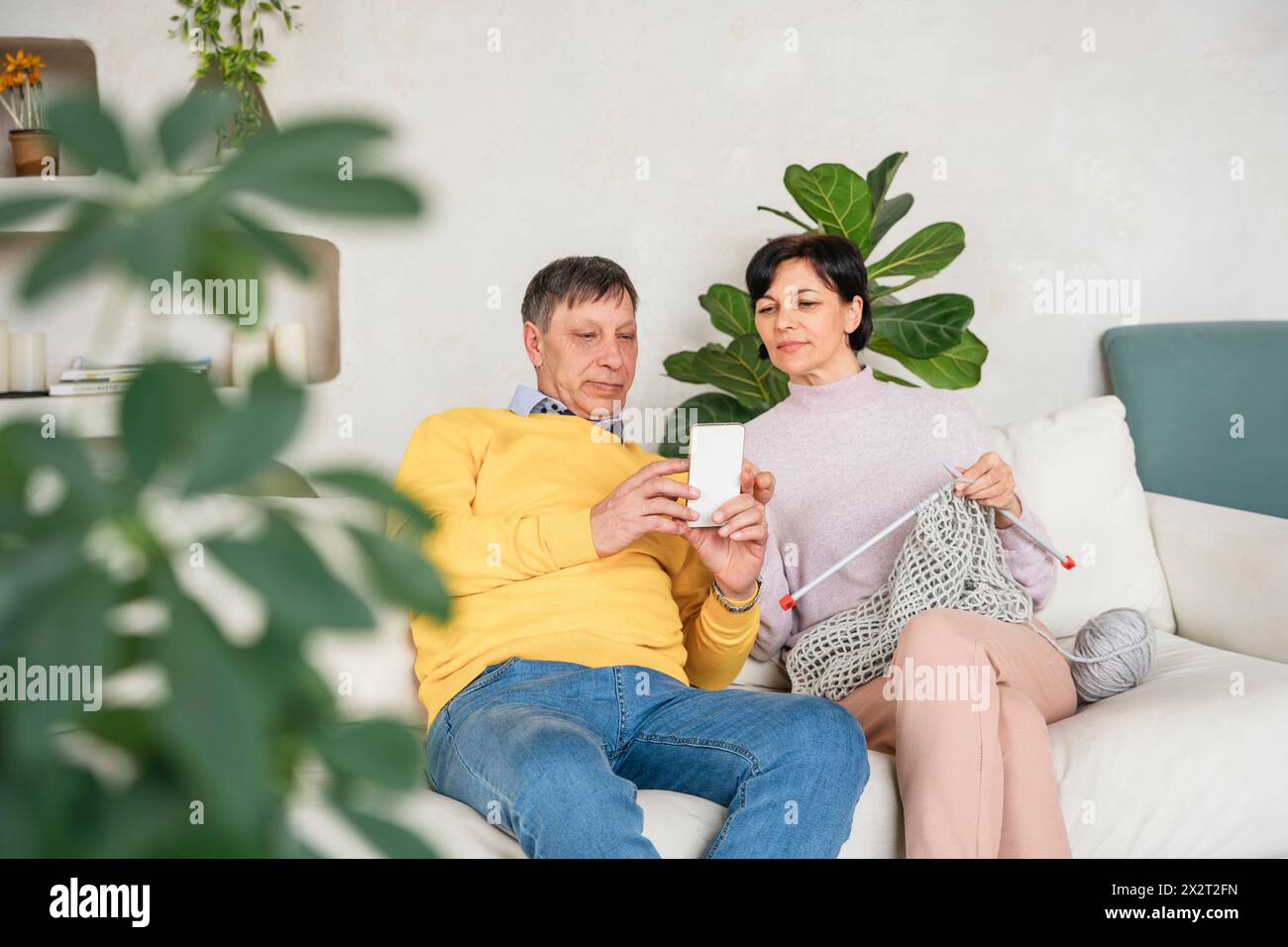 Mann zeigt Smartphone einer Frau, die zu Hause auf dem Sofa strickt Stockfoto