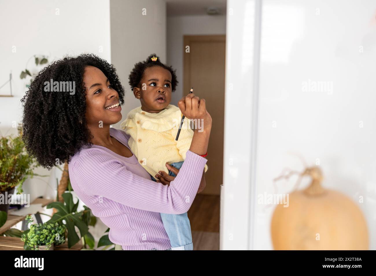 Lächelnde junge alleinerziehende Mutter, die Tochter trägt, die auf dem Kühlschrank in der Küche zu Hause schreibt Stockfoto
