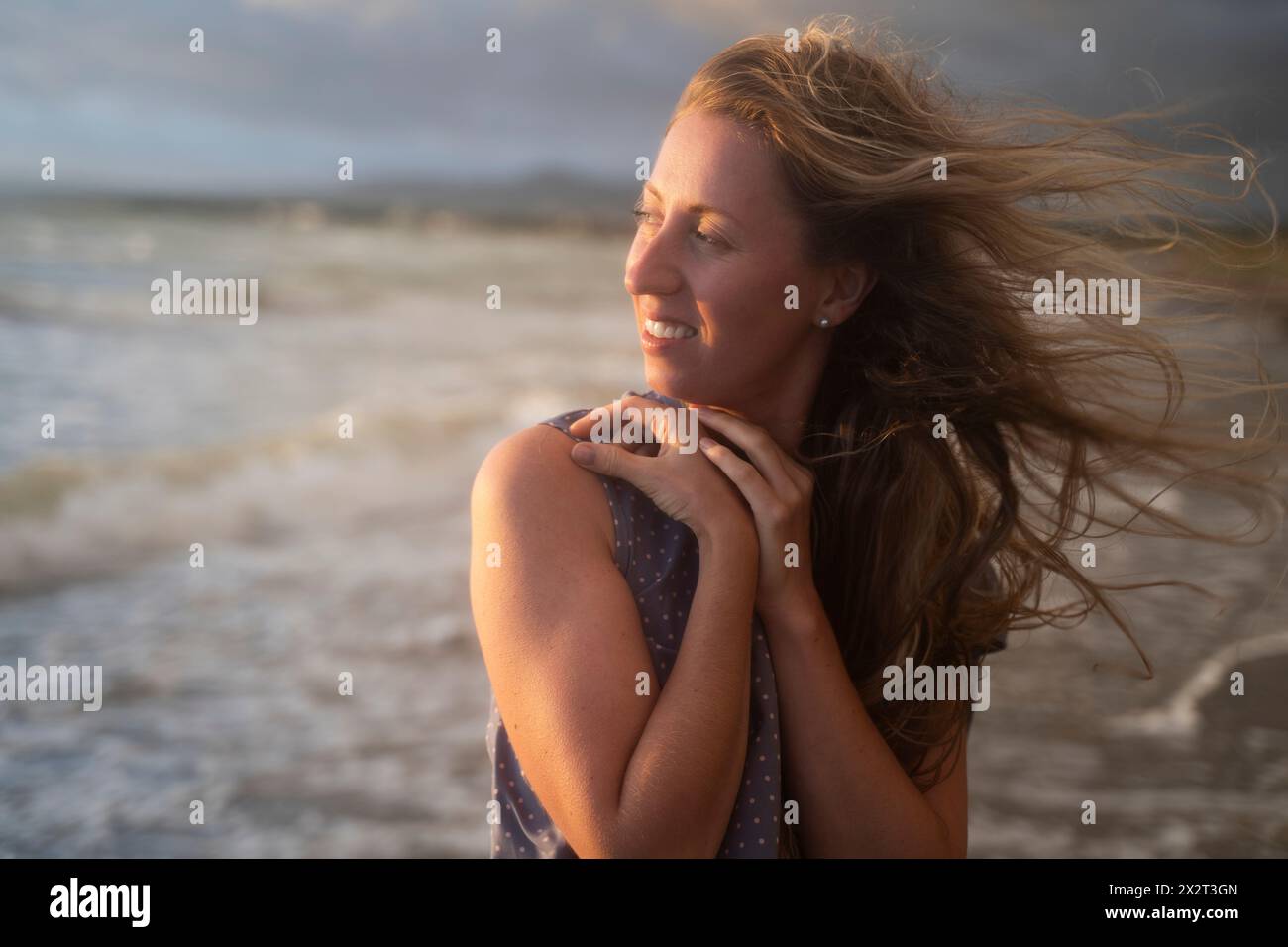 Lächelnde blonde Frau, die den Sonnenuntergang am windigen Strand genießt Stockfoto
