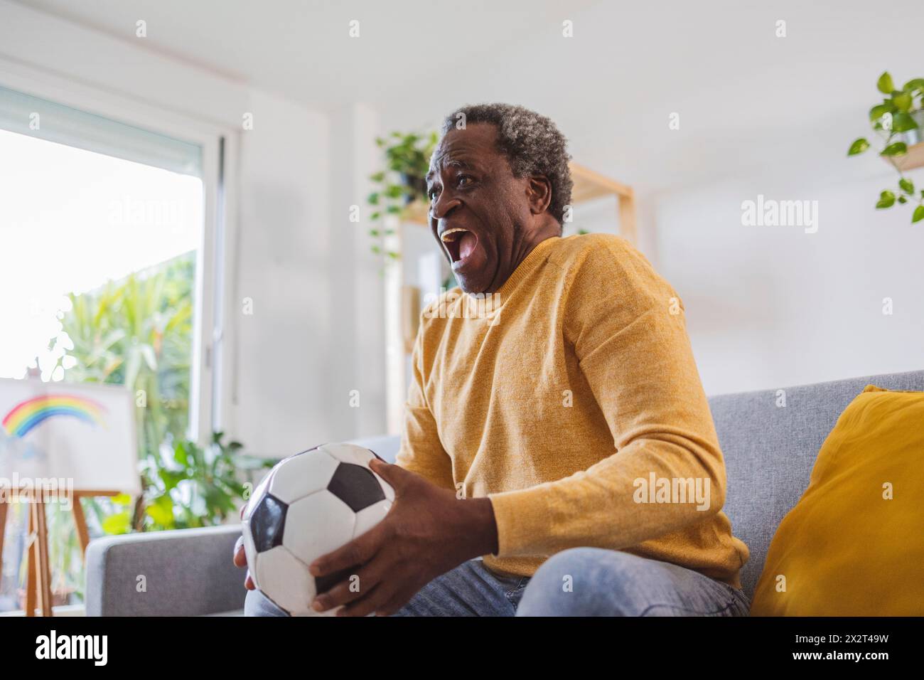 Glücklicher Senior-Mann, der Fußball hält und zuhause das Spiel ansieht Stockfoto