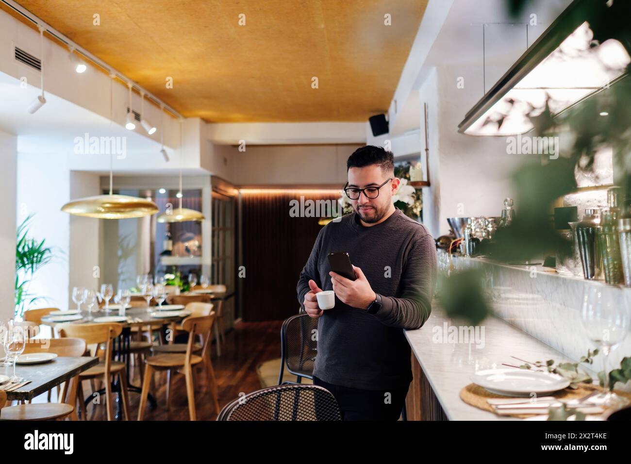 Kunde nutzt das Mobiltelefon, das sich an der Bar mit einer Kaffeetasse im Restaurant lehnt Stockfoto