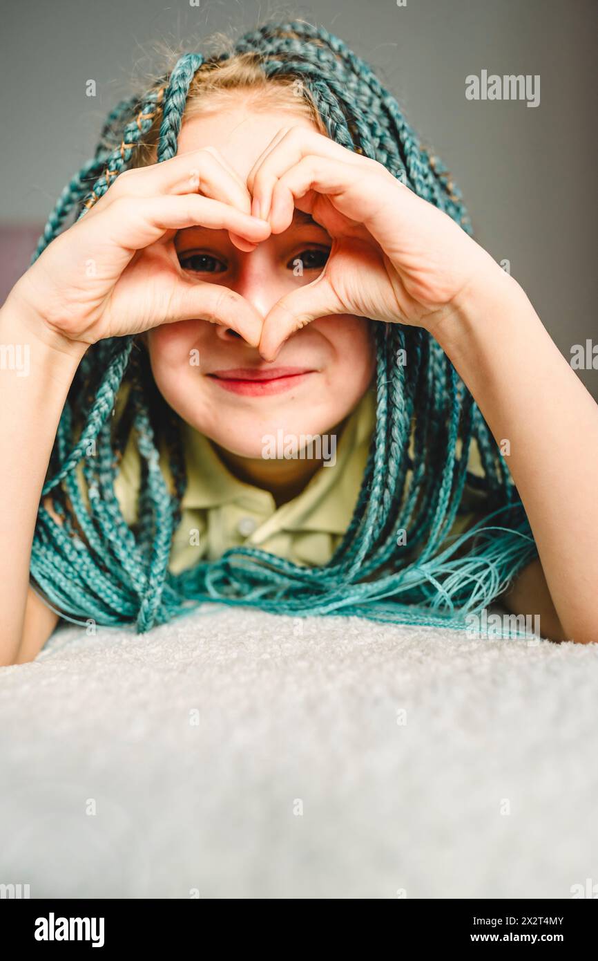 Lächelndes Mädchen mit türkisfarbenen geflochtenen Haaren, das auf dem Bett liegt und zu Hause Herzform macht Stockfoto