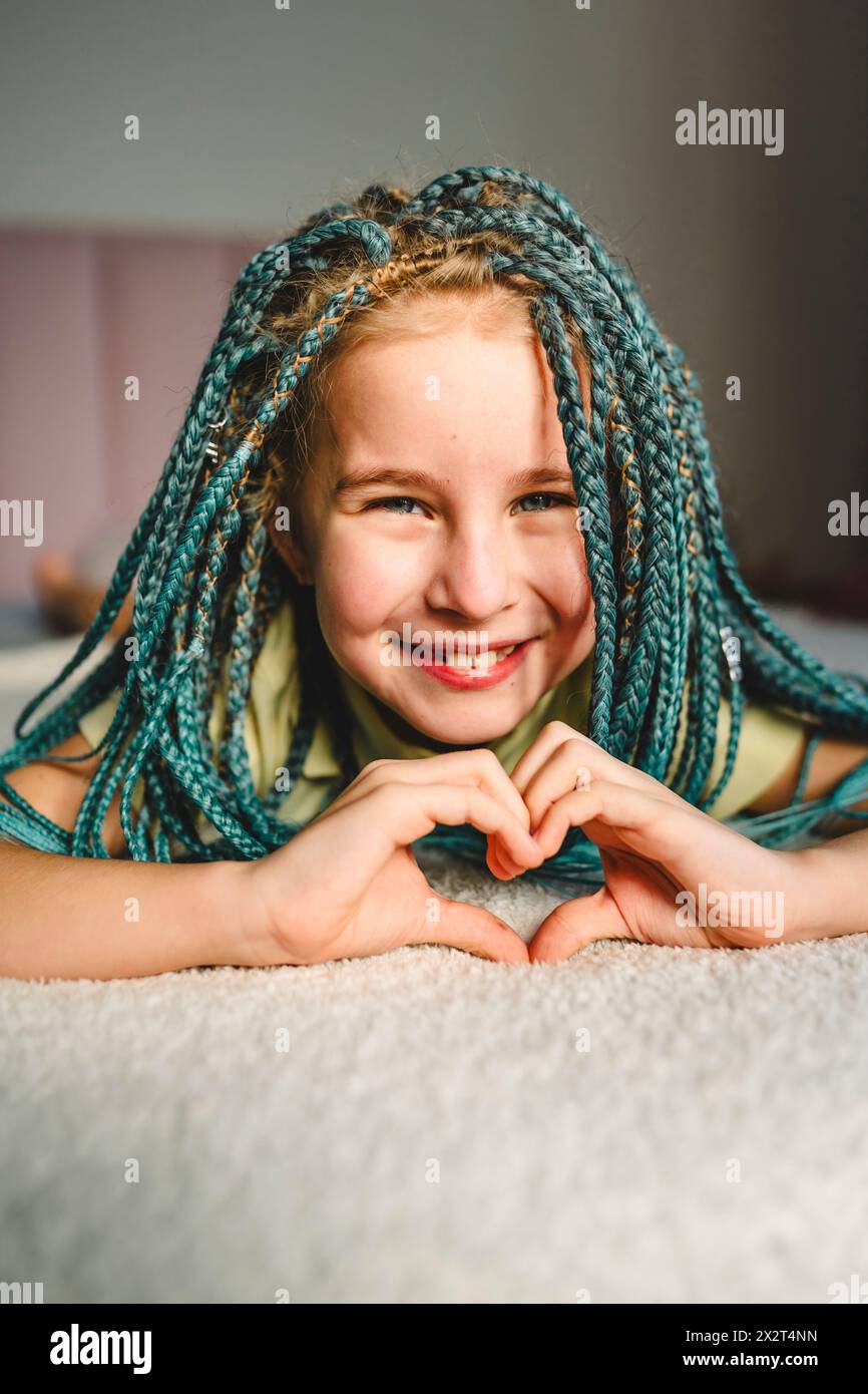 Glückliches Mädchen mit türkisfarbenen geflochtenen Haaren, das auf dem Bett liegt und zu Hause Herzform macht Stockfoto