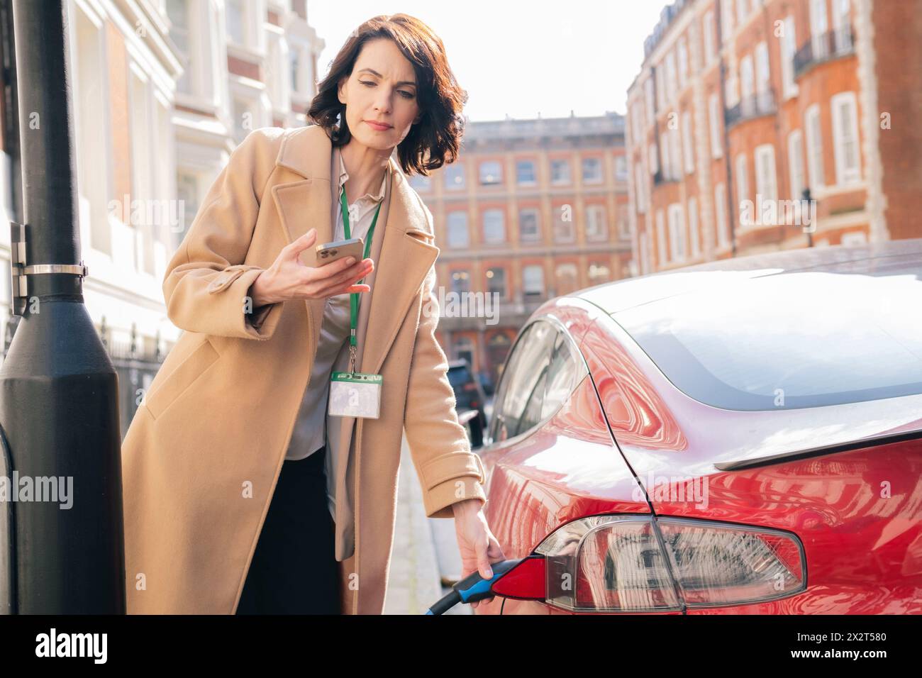 Geschäftsfrau, die in der Stadt ein Smartphone benutzt und ein Elektroauto auflädt Stockfoto