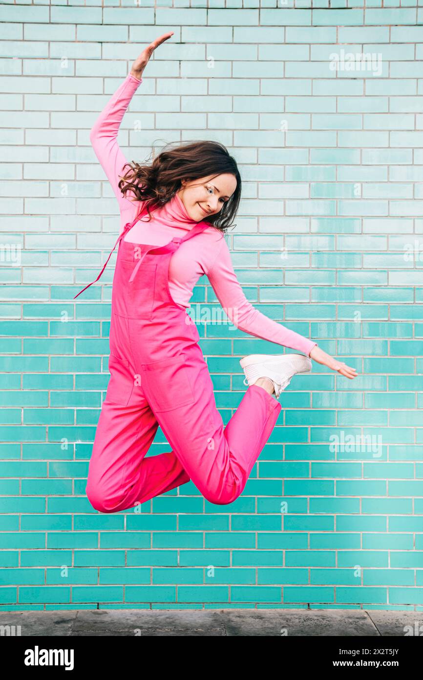 Glückliche Frau mit rosa Latzhose, die vor einer blauen Ziegelwand springt Stockfoto