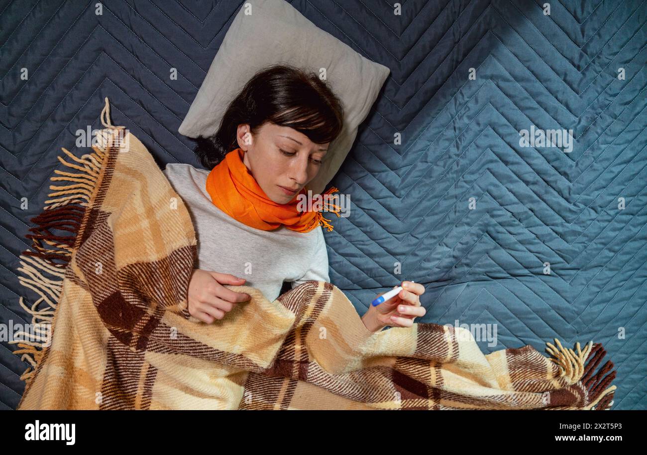 Frau mit Decke, die die Temperatur durch Thermometer auf dem Bett zu Hause überprüft Stockfoto