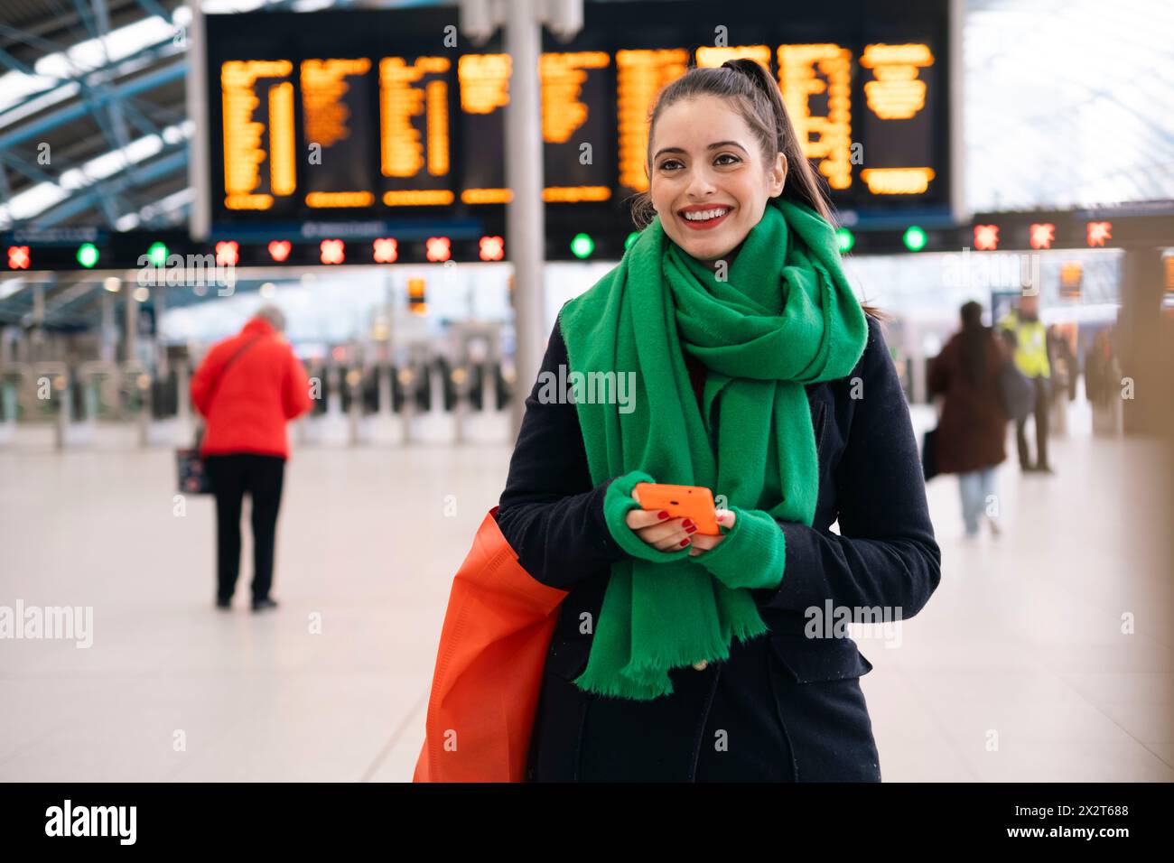 Glückliche Frau, die einen grünen Schal trägt und am Bahnhof ihr Smartphone hält Stockfoto
