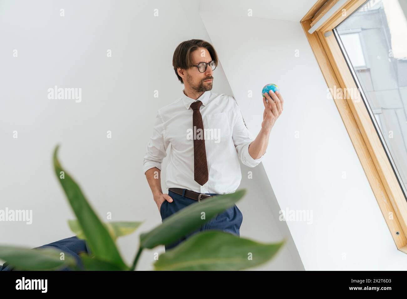 Selbstbewusster Geschäftsmann, der im Büro einen Globus hält und sich an eine weiße Wand lehnt Stockfoto