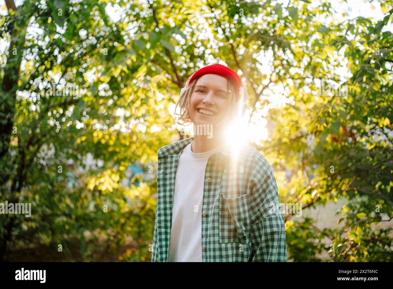 Lächelnde junge Frau mit Strickmütze und kariertem Hemd vor Bäumen im Park Stockfoto