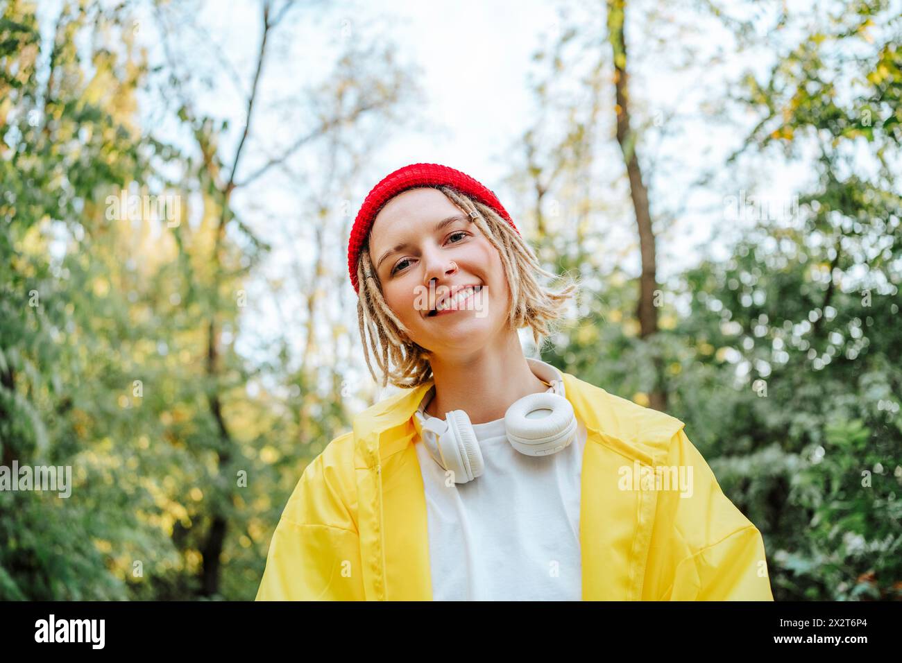 Lächelnde junge Frau mit Strickmütze und gelbem Regenmantel im Park Stockfoto