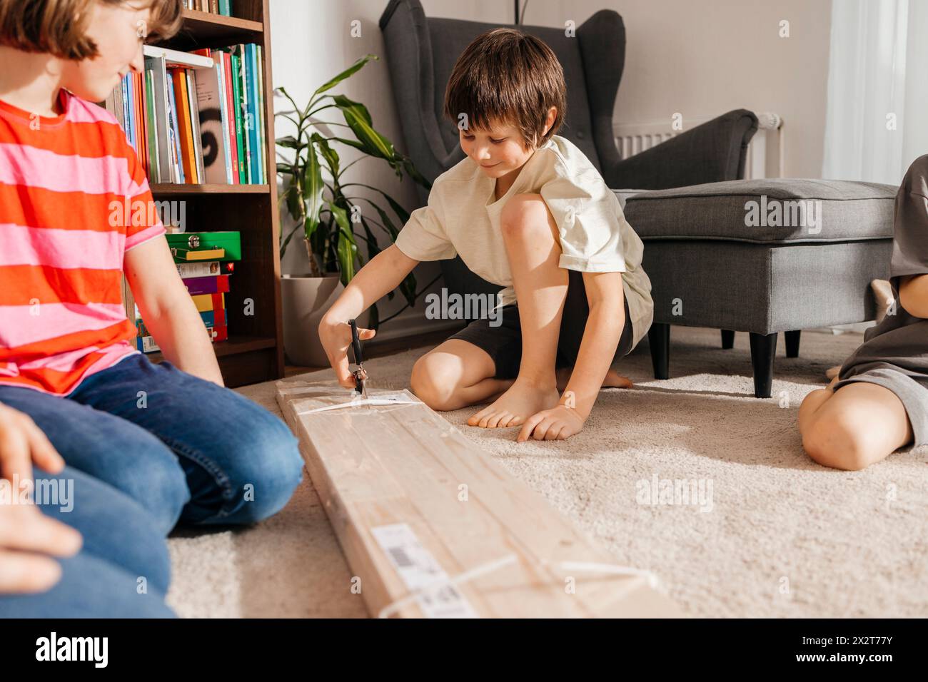Boy Opening Plank Paket mit Schere von Geschwistern und Vater kniend im Wohnzimmer Stockfoto