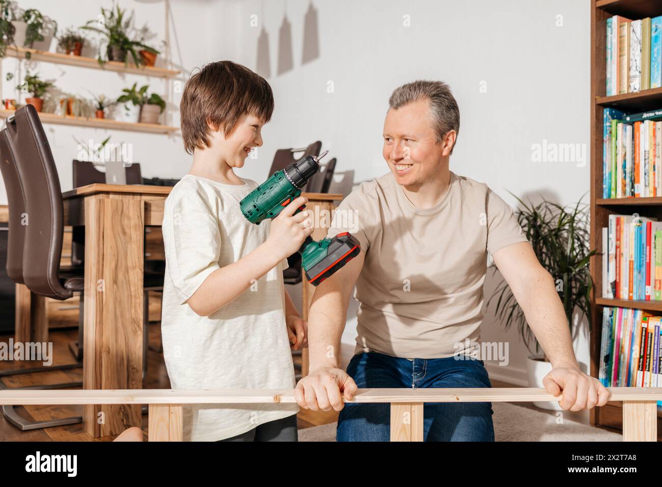 Lächelnder Junge, der Elektrowerkzeug hält, vom Vater, der Möbel zu Hause zusammenbaut Stockfoto