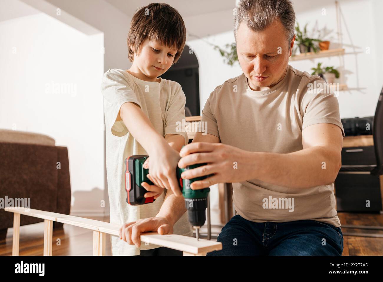Vater hilft dem Sohn beim Einsatz eines Elektrowerkzeugs für die Montage von Holzmöbeln Stockfoto