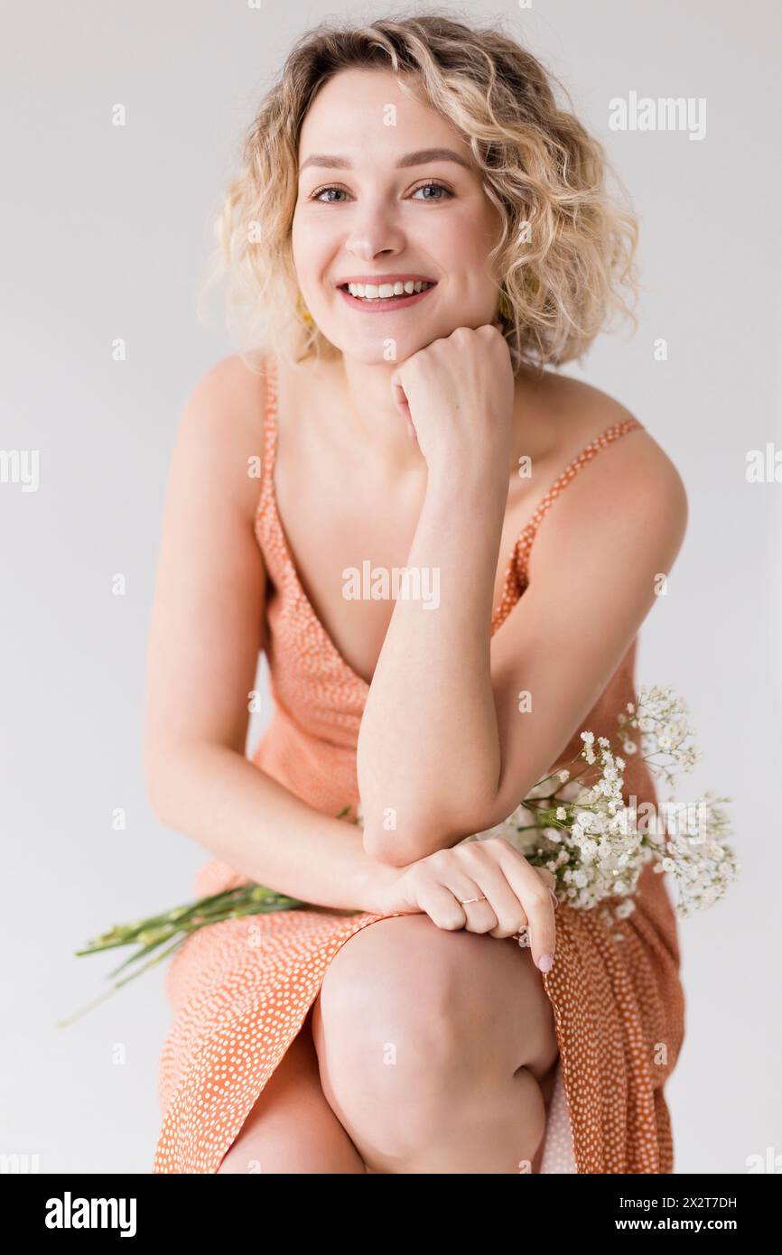 Glückliche schöne blonde Frau, die mit Blumen vor weißem Hintergrund kniet Stockfoto