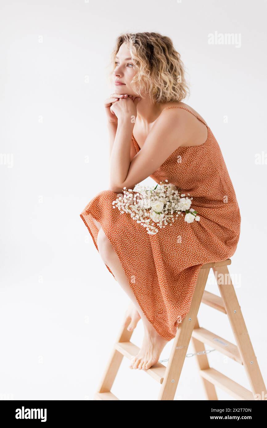 Nachdenkliche blonde Frau mit Blumen, die auf einer Leiter vor weißem Hintergrund sitzt Stockfoto