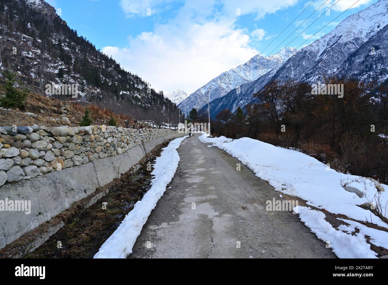Schneebedeckte Straße und Steinmauer in Rakcham, Himachal Pradesh, Indien, mit majestätischen Himalaya-Bergen im Hintergrund unter blauem Himmel. Stockfoto