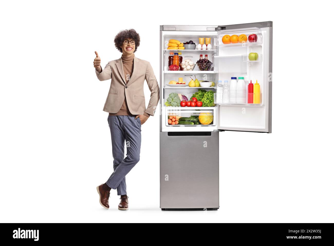 Glücklicher junger Mann, der Daumen nach oben drückt und neben einem Kühlschrank steht, isoliert auf weißem Hintergrund Stockfoto