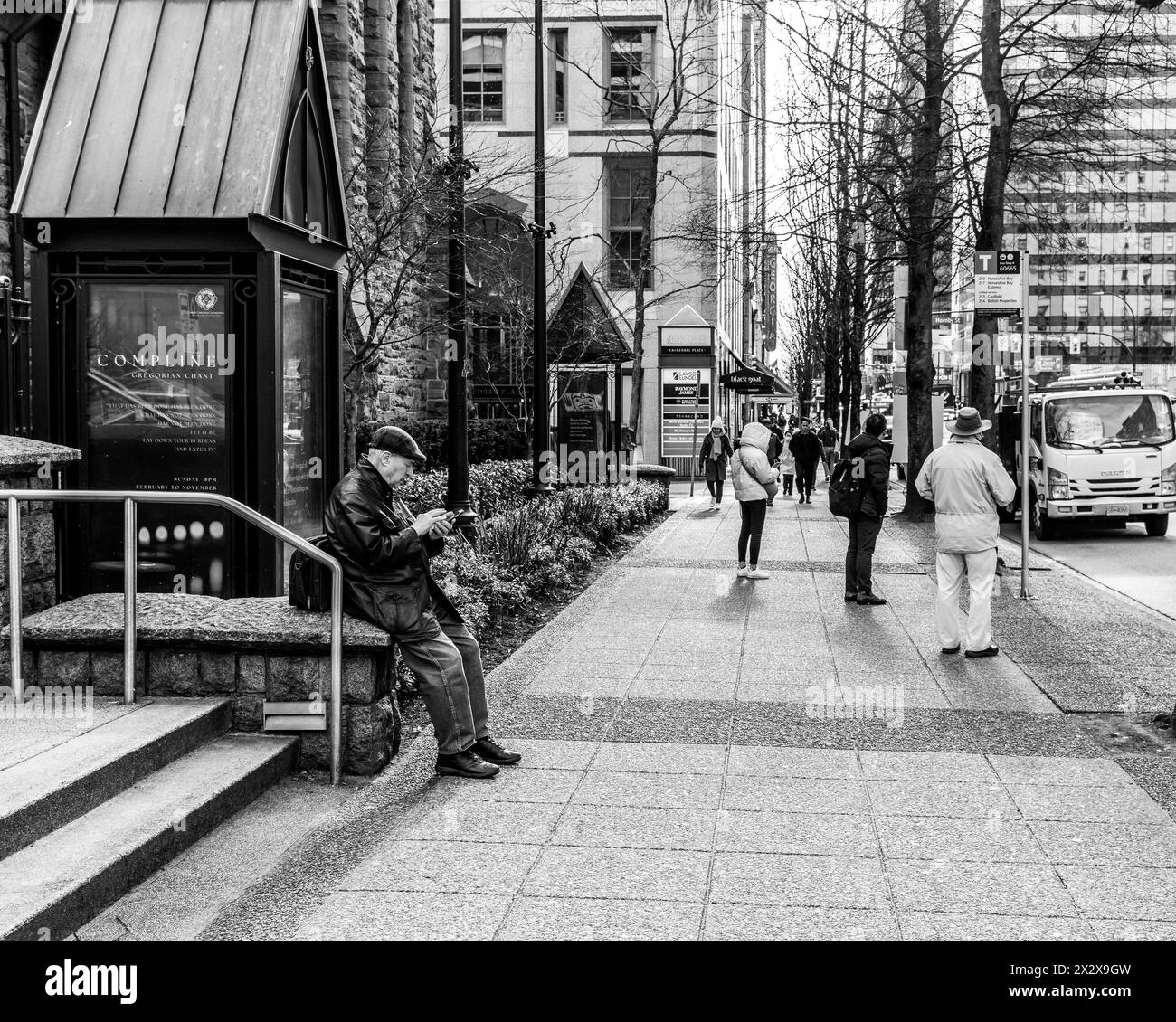 Vancouver, Kanada - 27. Februar 2024: Ein Schwarzweißfoto eines älteren Mannes in flacher Mütze, der auf sein Handy schaut, während er mit anderen an der Bushaltestelle wartete Stockfoto