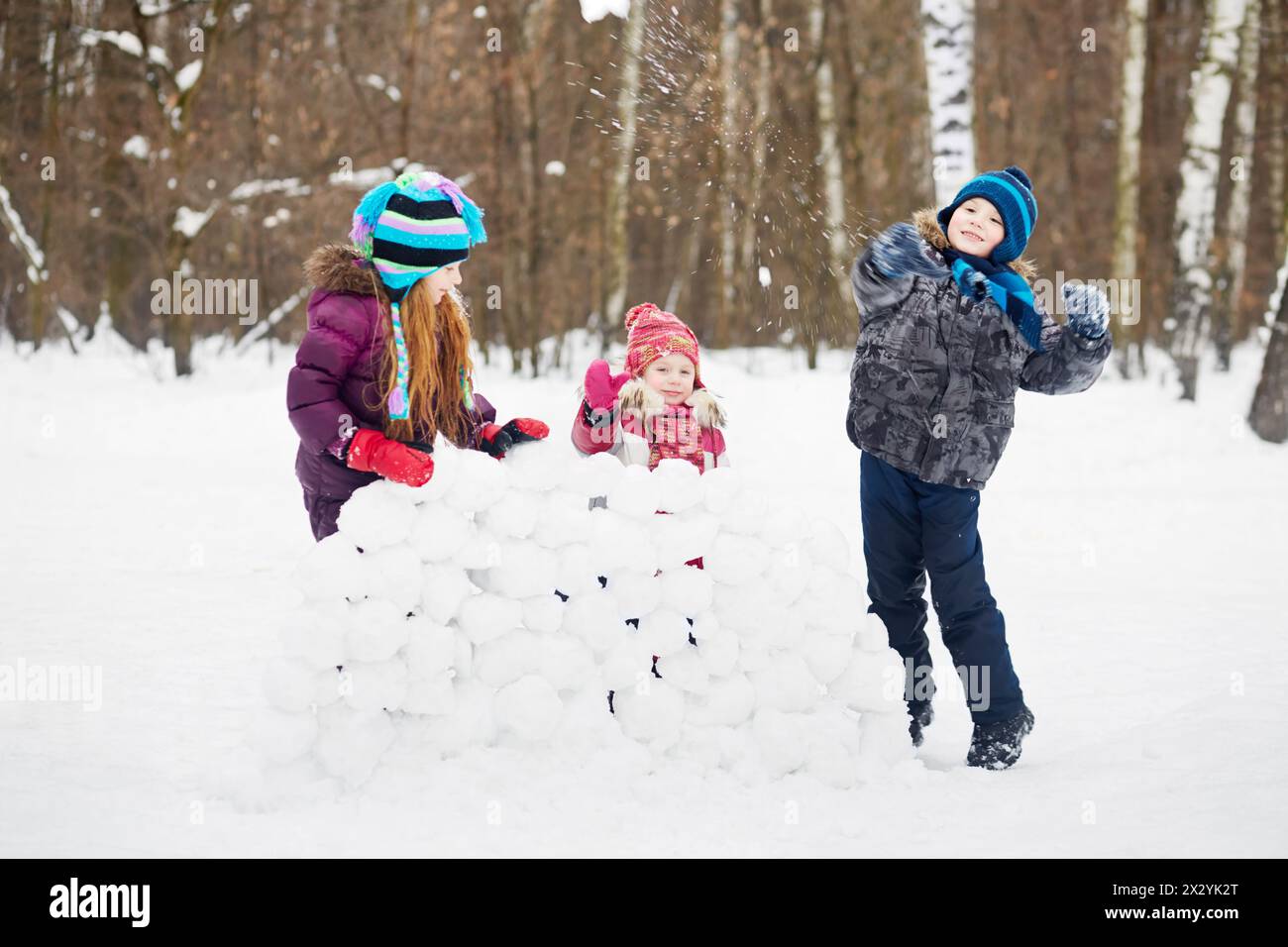 Zwei Mädchen stehen hinter einer Wand aus Schneeblöcken und ein Junge wirft Schneebälle Stockfoto