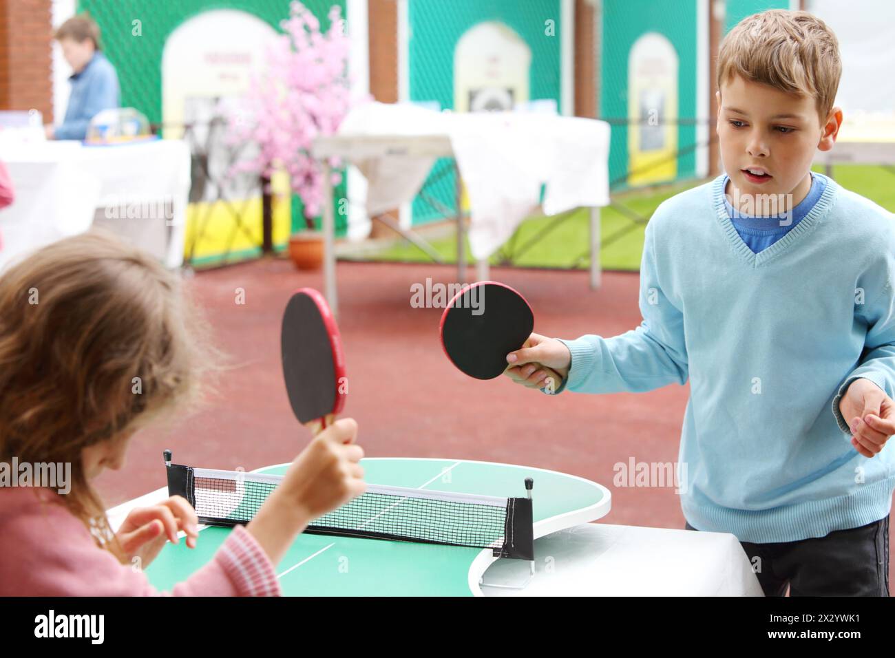 Kleines Mädchen und Junge in Blau spielen Tischtennis im Park am Sommertag. Konzentriere dich auf den Jungen. Stockfoto