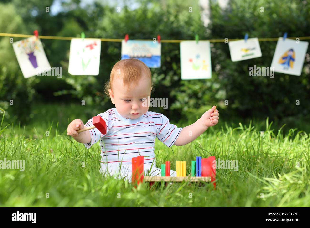 Ein kleiner Junge spielt mit bunten Holzspielzeugen auf dem Gras, im Hintergrund hängen Kinderzeichnungen Stockfoto