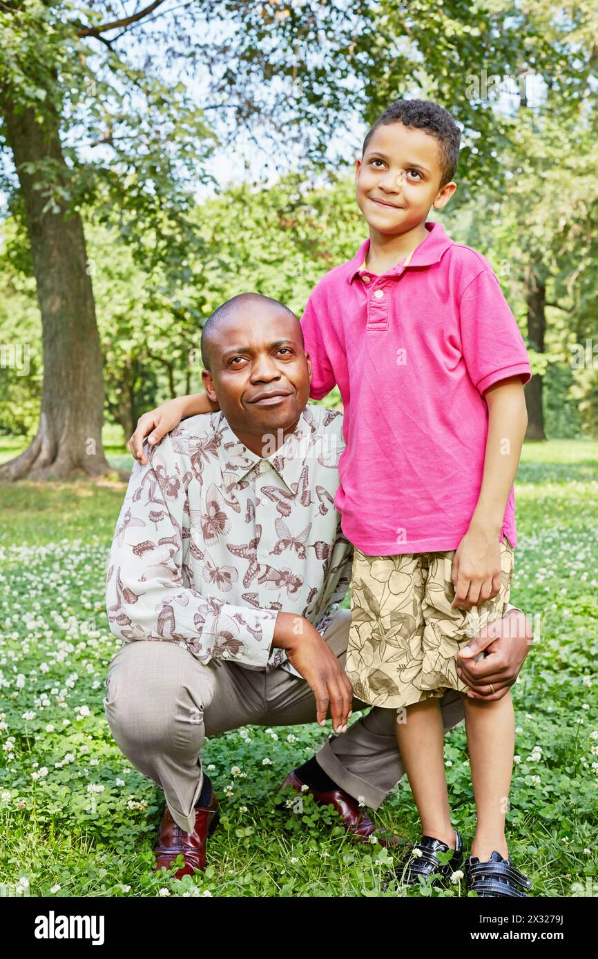 Der afrikanische Vater sitzt in der Hocke und sein kleiner Sohn steht in der Nähe und hält die Hand auf den Schultern des Vaters Stockfoto