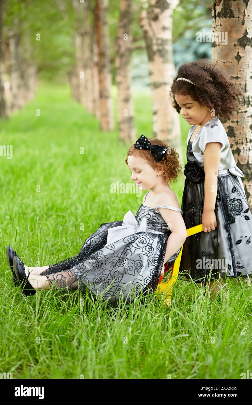 Das kleine Mädchen trägt ein schönes schwarzes Kleid und trägt ein anderes Mädchen auf einer Plastikkarre auf dem Rasen im Park Stockfoto