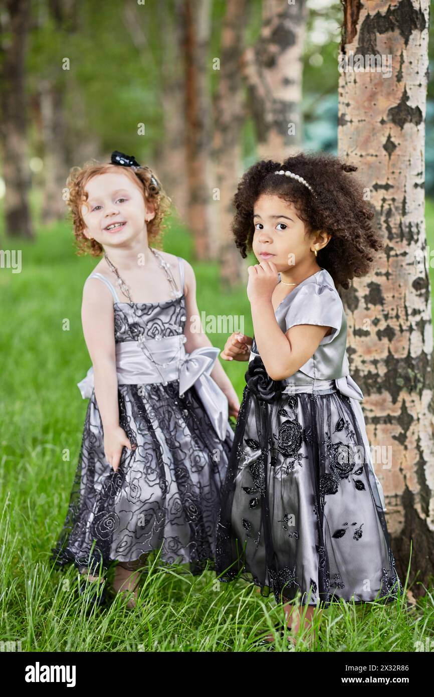 Zwei kleine Mädchen in wunderschönen schwarzen Gewändern stehen auf dem grasbewachsenen Rasen in der Nähe eines Baumes im Park und konzentrieren sich auf Mulatten-Mädchen Stockfoto