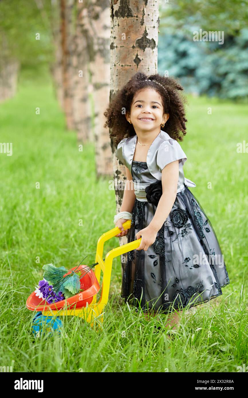 Ein lächelndes kleines Mädchen in einem wunderschönen schwarzen Kleid steht im Park und hält eine Plastikkarre, in der Blumen liegen Stockfoto