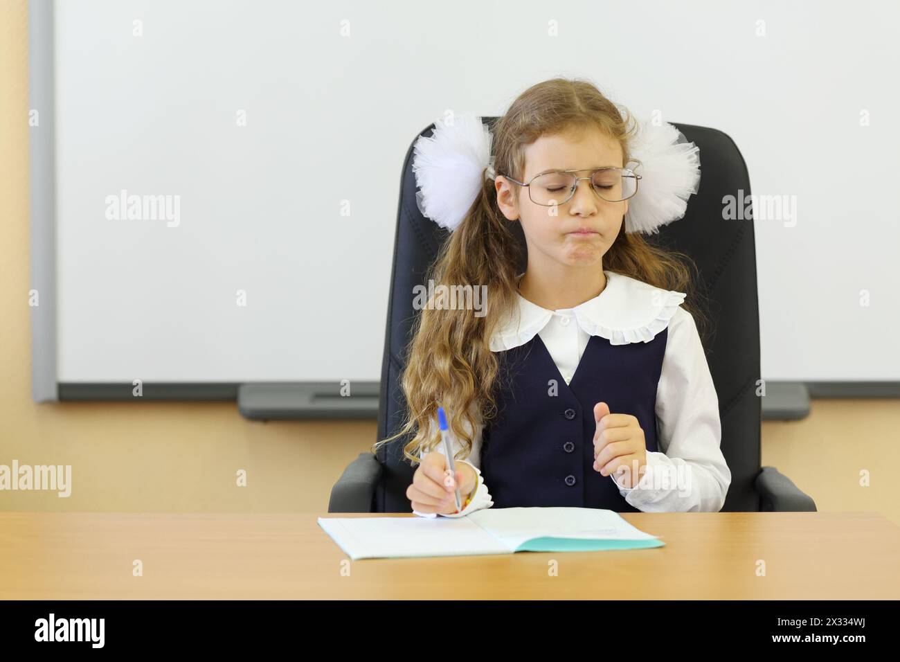 Mädchen mit Brille sitzt am Lehrertisch mit Übungsbuch im Klassenzimmer und denkt in der Schule. Stockfoto