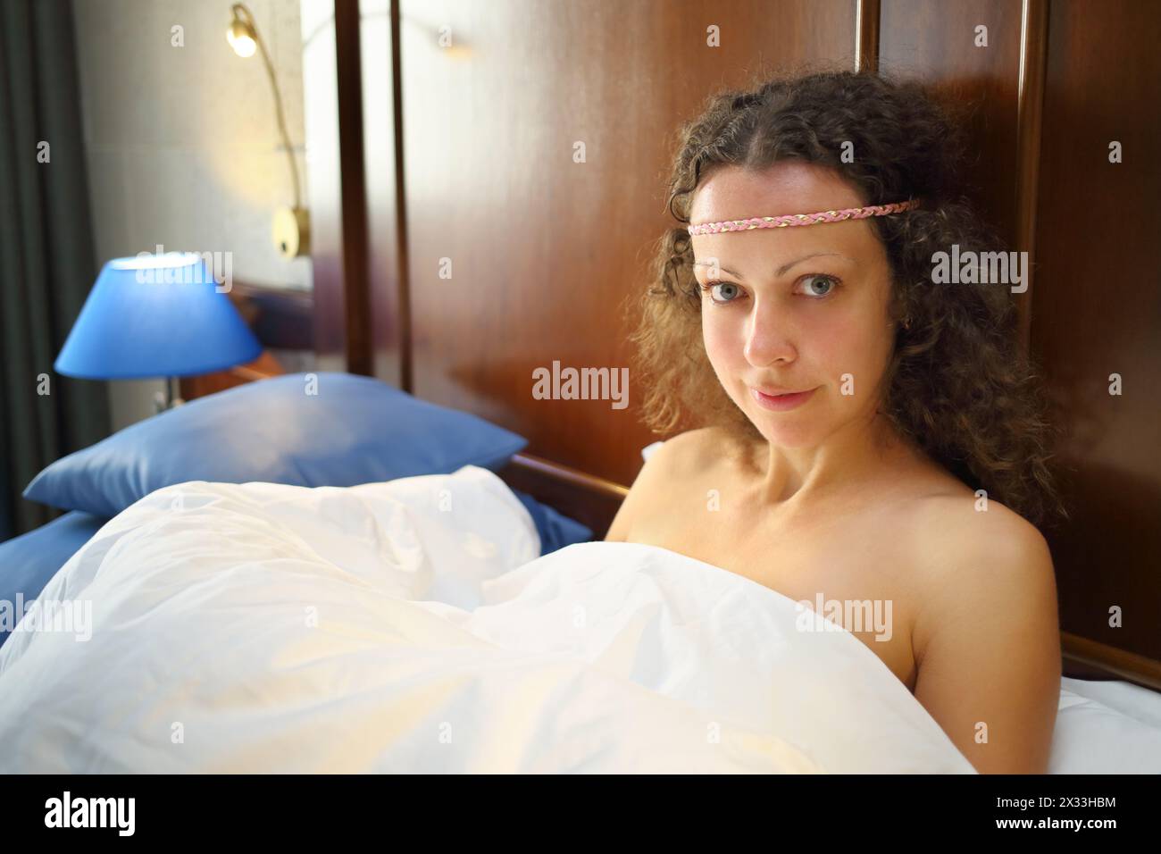 Porträt einer schönen Frau mit lockigem Haar unter einer weißen Decke im Bett Stockfoto