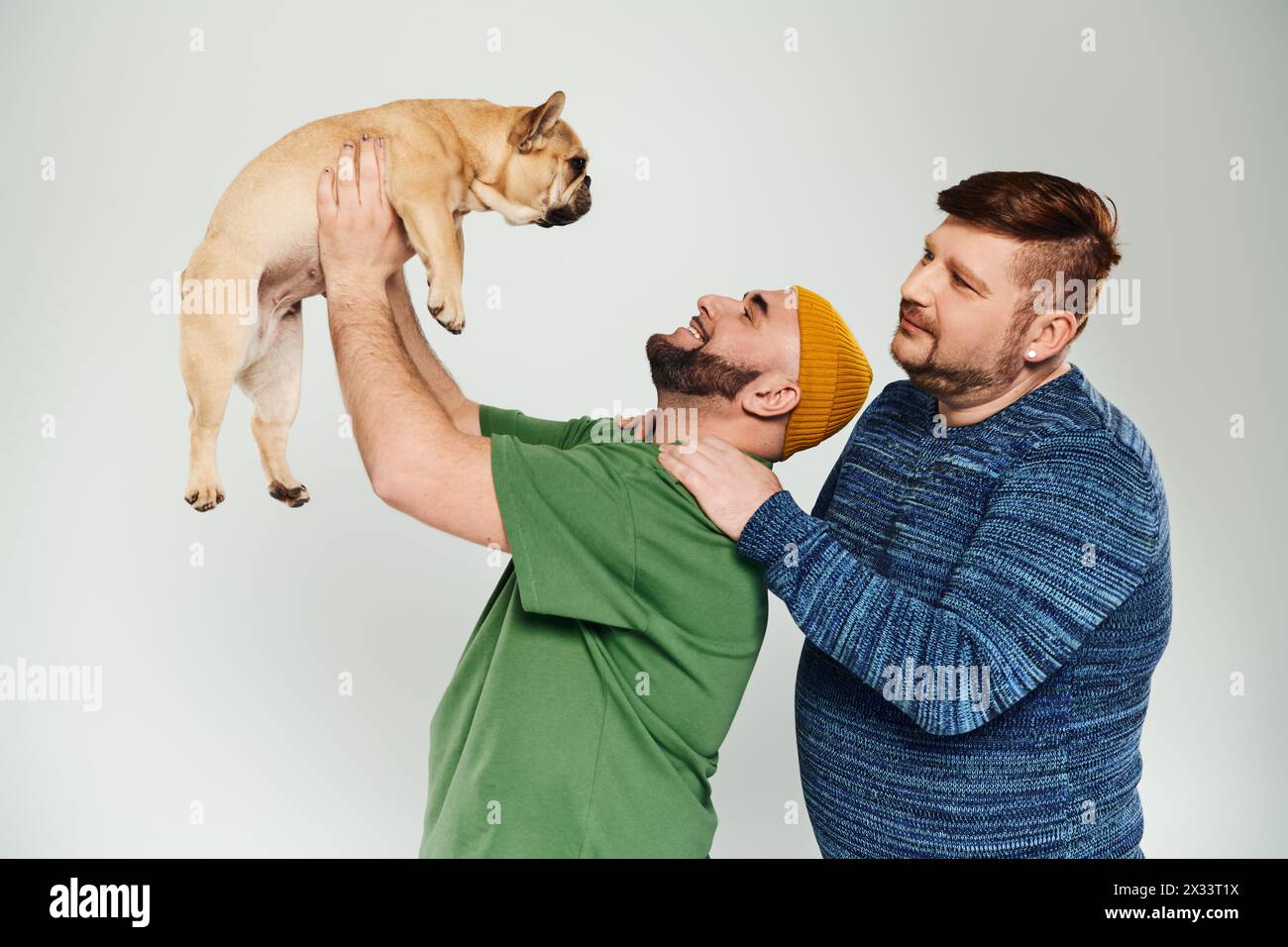 Zwei Männer halten eine französische Bulldogge in einem zärtlichen Moment zart nah an sein Gesicht. Stockfoto