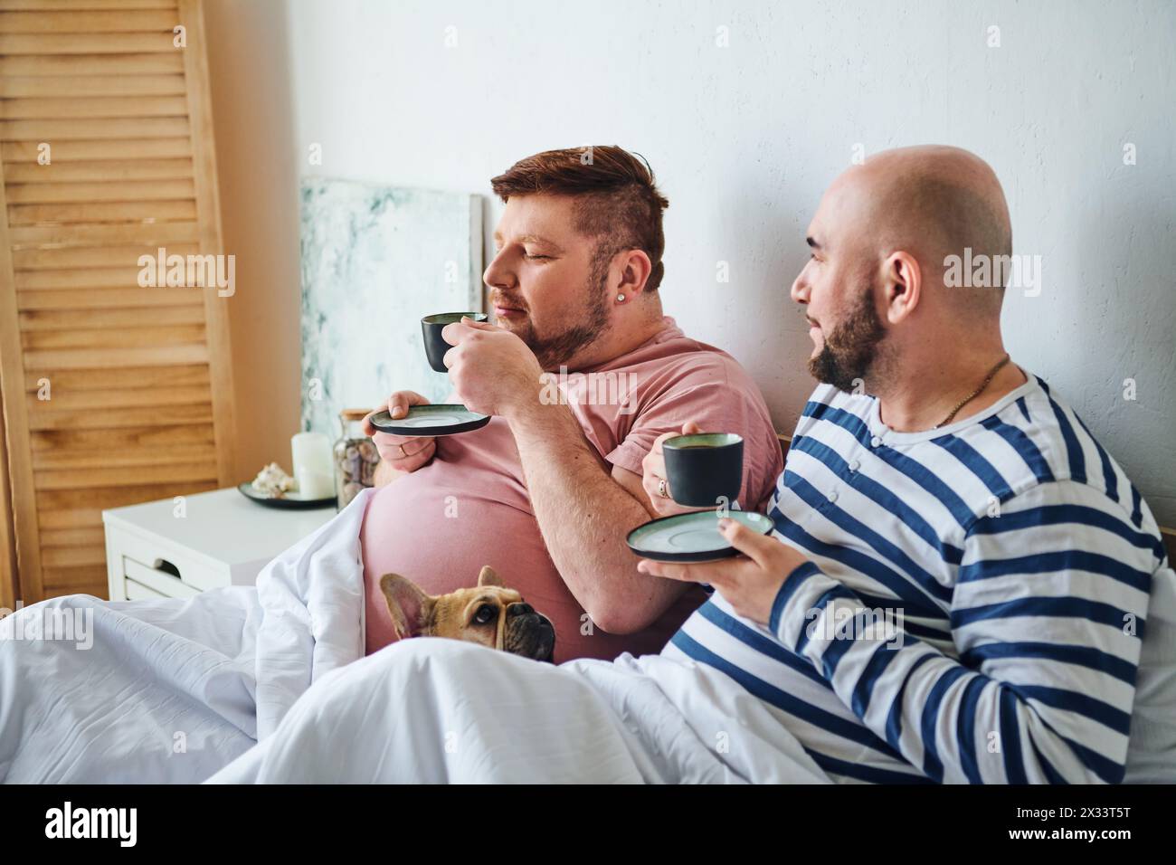 Ein schwules Paar und ihre französische Bulldogge liegen auf einem Bett. Stockfoto