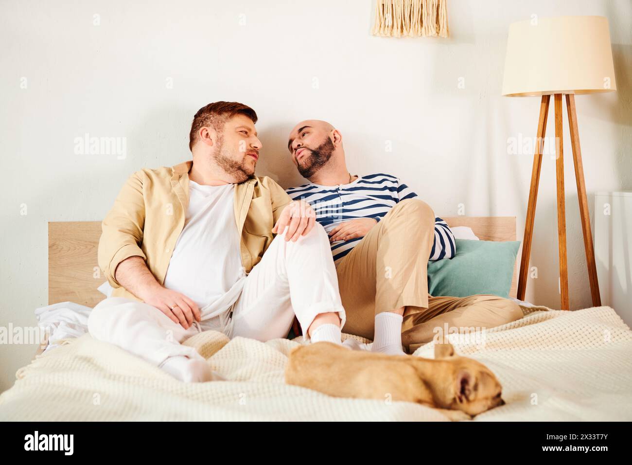 Ein attraktives schwules Paar sitzt bequem auf einem Bett mit ihrer französischen Bulldogge. Stockfoto