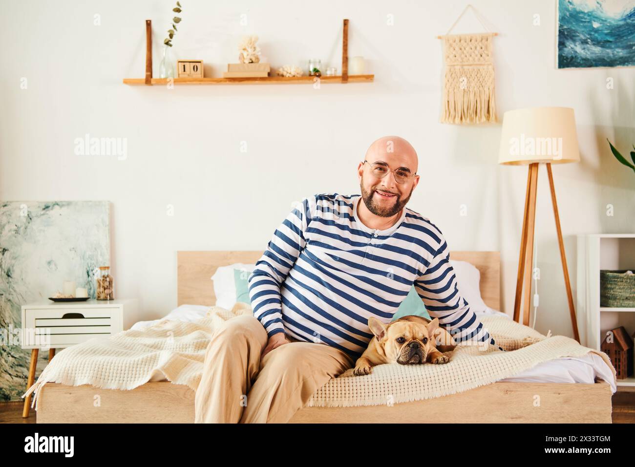 Ein Mann mit Brille sitzt friedlich auf einem Bett, begleitet von seiner französischen Bulldogge. Stockfoto