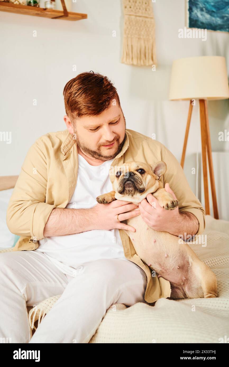 Ein Mann, der auf einem Bett sitzt und liebevoll eine kleine französische Bulldogge hält. Stockfoto
