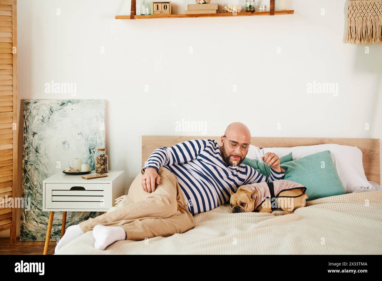 Hübscher Mann mit Brille auf einem Bett neben einer französischen Bulldogge. Stockfoto