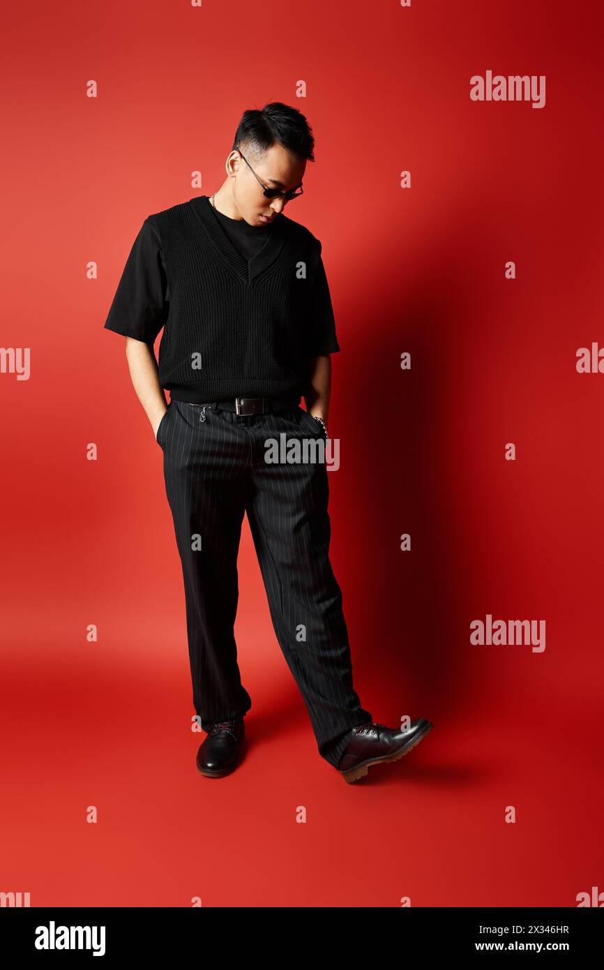 Ein stilvoller und hübscher asiatischer Mann in schwarzer Kleidung posiert selbstbewusst vor einem leuchtend roten Hintergrund in einem Studio. Stockfoto