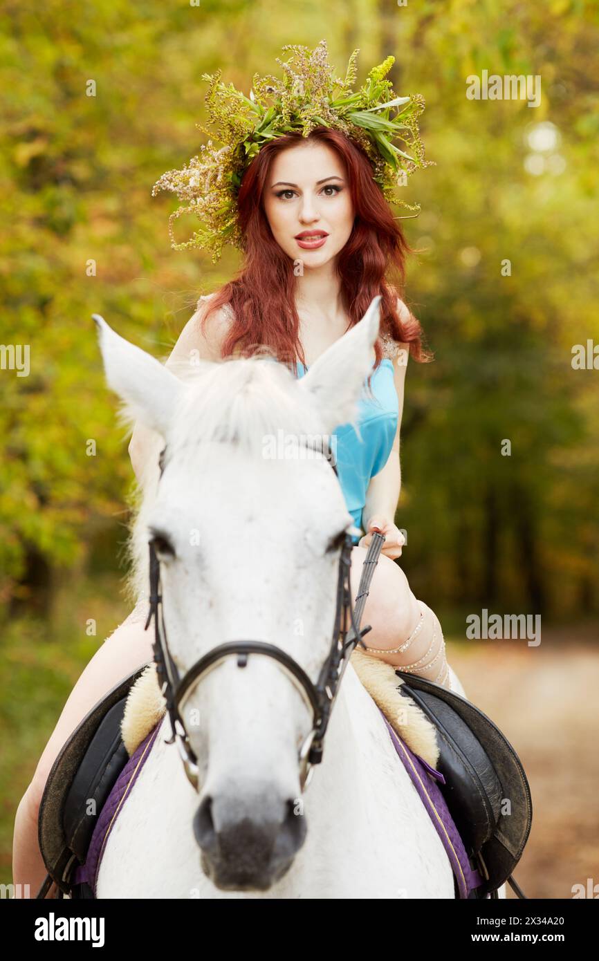 Rothaarige Frau mit Blumenkranz auf Kopfritten auf weißem Pferd im Park. Stockfoto