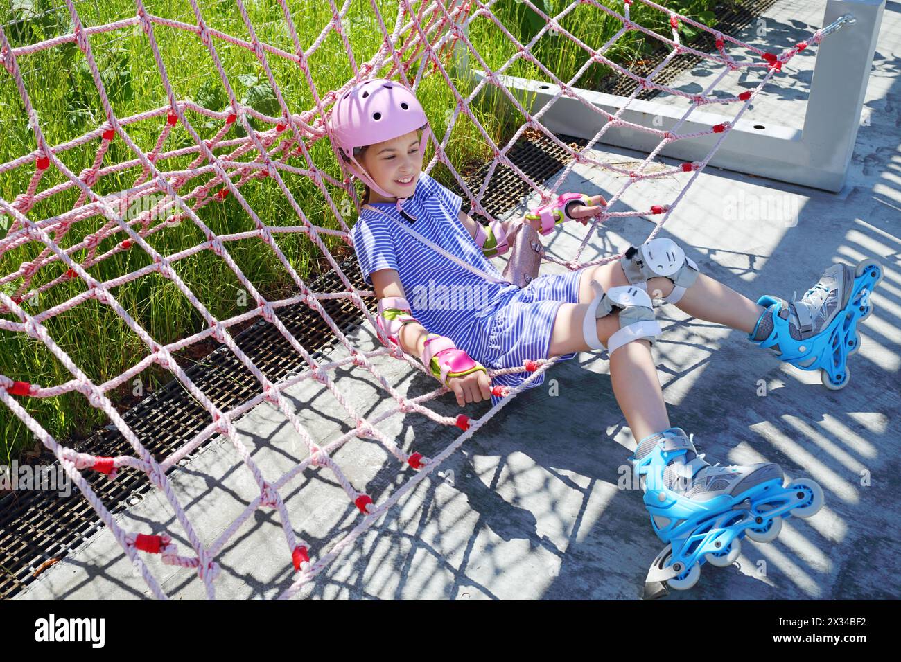 Mädchen mit rosa Helm und Rollen sitzt im Sommer in einer Netzhängematte im Skatepark Stockfoto