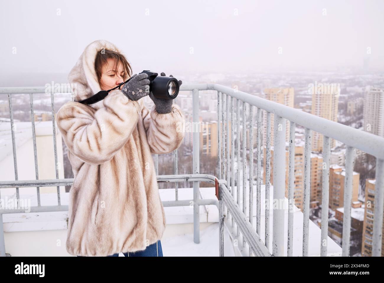 Frau in Pelzjacke macht Fotos von der Stadt, die im Winter auf dem Dach des Gebäudes steht. Stockfoto