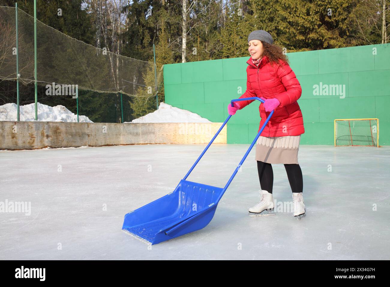 Glückliche Frau in einer roten Jacke mit einem großen Spaten auf einer Eislaufbahn Stockfoto