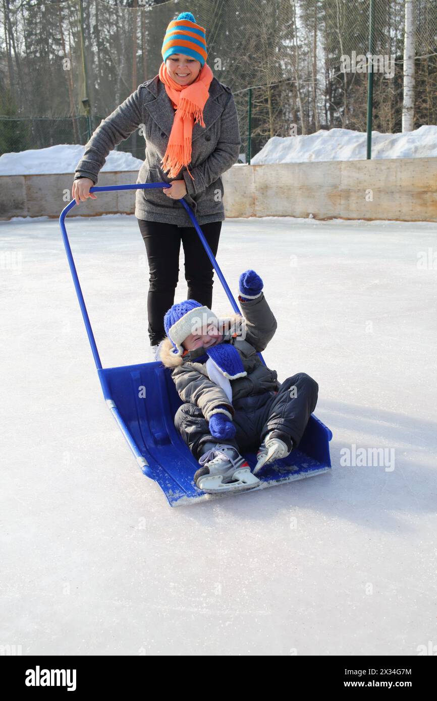 Mutter rollt den kleinen Sohn in einer großen Schneeschaufel auf einer Eislaufbahn Stockfoto