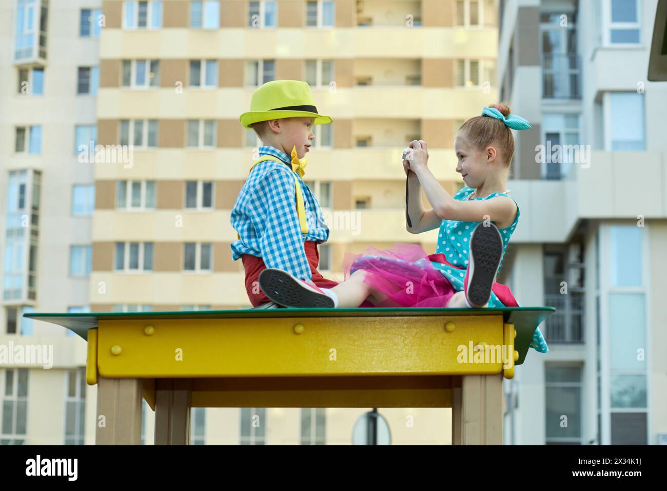 Das kleine Mädchen fotografiert den kleinen Jungen, der auf einem Holzdach auf dem Spielplatz sitzt. Stockfoto