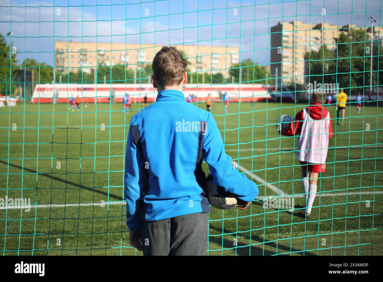 Junger Mann in einer blauen Sportjacke mit einem Ball, der ein Fußballspiel im Stadion über dem Netz beobachtet, Blick von hinten Stockfoto