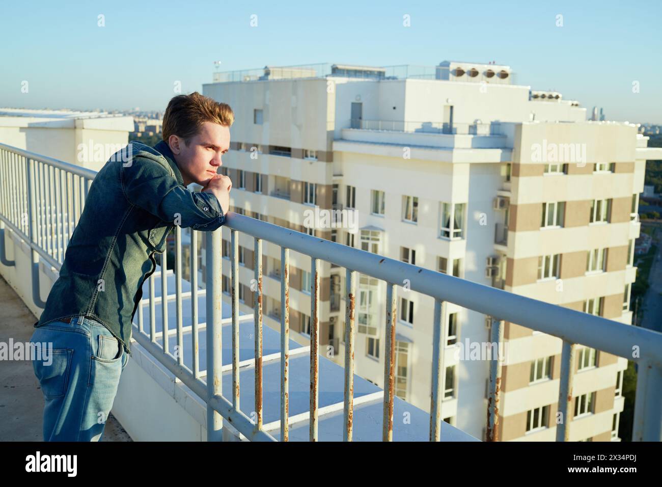 Ein junger Mann in Denim-Kleidung steht auf dem Dach des Gebäudes, der zum Zaun geneigt ist. Stockfoto