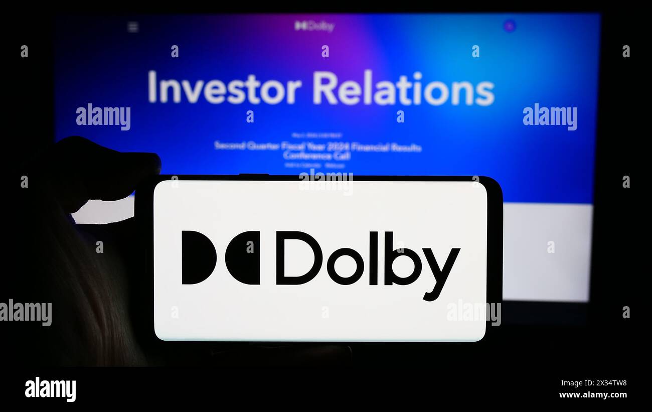 Person, die ein Mobiltelefon mit dem Logo des Audiotechnologieunternehmens Dolby Laboratories Inc. Vor der Webseite des Unternehmens hält. Schwerpunkt auf dem Display des Telefons. Stockfoto