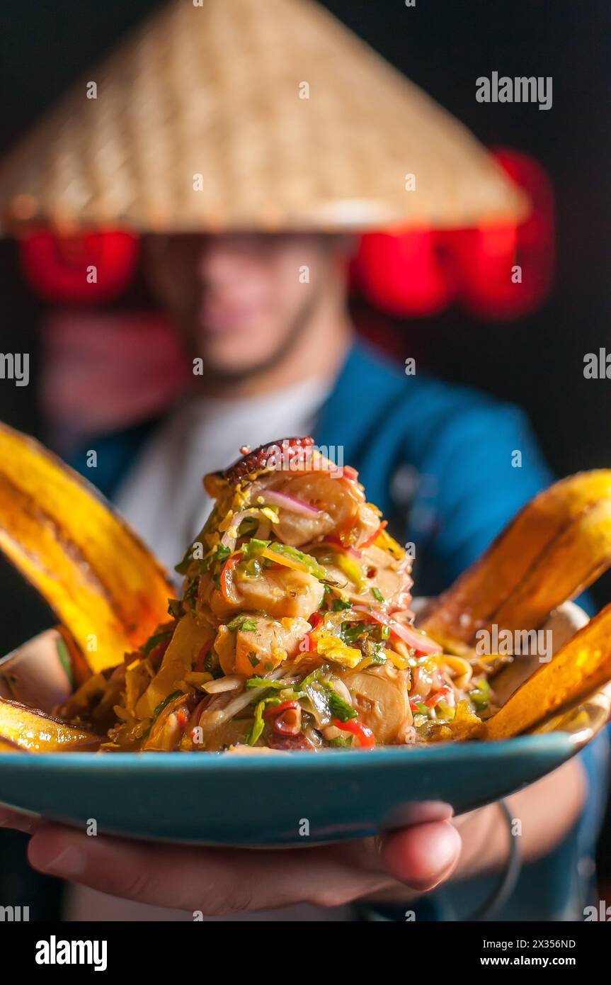 Ein Typ in japanischer Verkleidung und konischer Hut, der einen Teller Ceviche nikkei mit roten Laternen hält und serviert Stockfoto