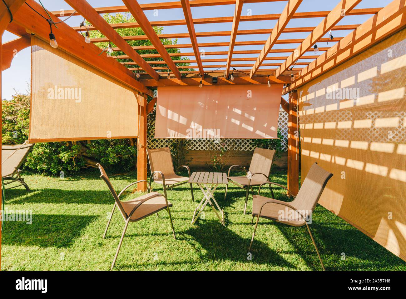 Pergola aus Holz mit Gartenmöbeln und Hängelampen in einem Garten, der Schatten auf den Boden wirft. Stockfoto