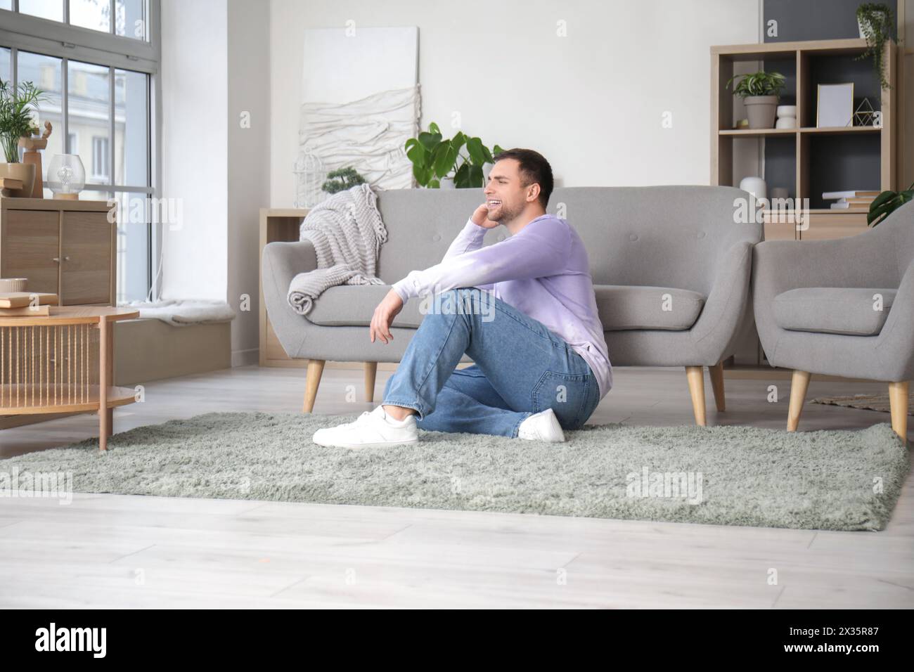 Junger Mann, der zu Hause neben dem grauen Sofa sitzt Stockfoto